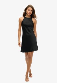 Kobieta idąca w czarnej halterowej sukience bez rękawów i przezroczystych obcasach na białym tle.