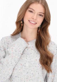 Világosszürke pulóver színes foltokkal, bordázott nyakkal és mandzsettákkal. Hosszú barna haj és csillag alakú fülbevalók egészítik ki a megjelenést.