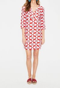 Rotes Blumenkleid mit weißen Hibiskusmustern, V-Ausschnitt, 3/4-Ärmel und knielange Schnittlänge. Kombiniert mit roten Rutschsandalen.