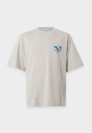 Jeune homme debout, les mains dans les poches, portant un t-shirt beige et un jogging assorti avec des logos bleus et des baskets blanches.