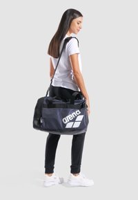 Bolsa de deporte negra con el logo blanco de "arena", que cuenta con un bolsillo de malla y dobles asas, llevada al hombro por una persona con ropa casual.