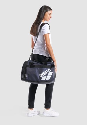 Borsa a tracolla nera con logo bianco "arena", dotata di tasca in rete e doppi manici, portata a spalla da una persona in abbigliamento casual.