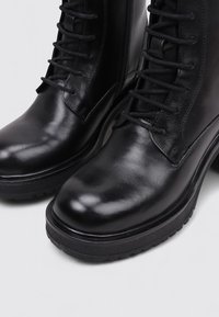 Bottes noires en cuir à lacets avec une finition lisse, hauteur élevée et semelle en caoutchouc durable. Les caractéristiques comprennent des coutures marquées et une fermeture éclair sur le côté.