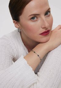 Srebrna bransoletka z owalnymi zielonymi akcentami noszona na jasnej skórze ramienia. Modelka w teksturowanym białym swetrze i kolczykach.