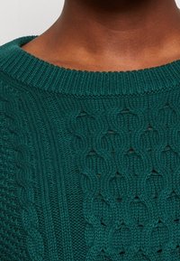 Pull vert en tricot avec un motif texturé comprenant des torsades et des mailles ajourées, col rond côtelé, et épaisseur moyenne.