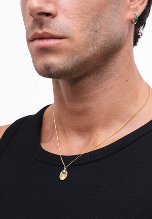 Vyras dėvi juodą marškinėlį be rankovių, auksinę grandinėlę su auksiniu ir mėlynu apskritu pakabuku bei mažą žalią lankinį auskarą kairėje ausyje.