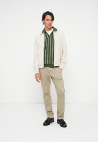 Jasno beżowa kurtka z zamkiem błyskawicznym na ciemnozielonej koszulce w paski, zestawiona z khaki spodniami i czarnymi loafersami, na jednolitym tle.