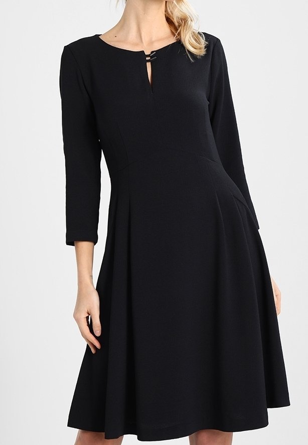 Femme portant une robe noire jusqu'aux genoux, à manches trois-quarts, avec un petit décolleté en encoche et deux fines bretelles horizontales.