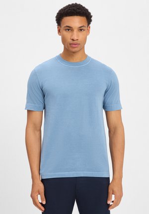 Mann trägt hellblaues T-Shirt mit kurzen Ärmeln und rundem Halsausschnitt sowie dunkle Hose, steht vor weißem Hintergrund.