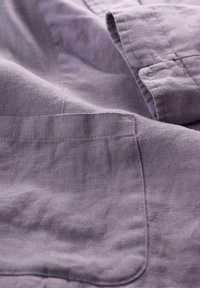 Tissu en lin violet clair avec une texture subtile et un détail de poche visible. Coutures surpiquées et tombé doux, mettant l'accent sur la qualité du tissu.