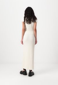 Rochie lungă tricotată în alb murdar, cu mâneci scurte, model texturat, siluetă ajustată și tiv fendu. Este asortată cu pantofi negri cu talpă groasă.