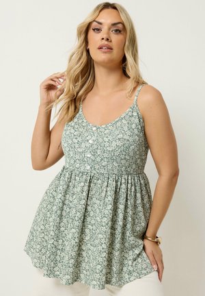 Kvinde med langt blondt hår iført en grøn blomsterprint ærmeløs kjole med knapper, poserer mod en ensfarvet lys baggrund.