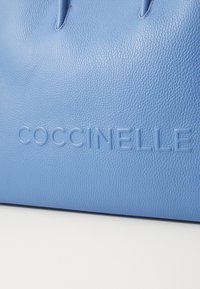 Sac à main en cuir bleu clair avec surface texturée, arborant le nom de la marque "Coccinelle" en relief sur le devant. Design minimaliste.