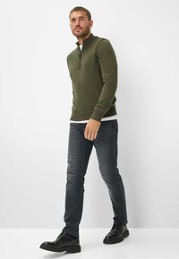 Pull en maille vert foncé avec un col zippé moyen, associé à un jean gris slim et des chaussures noires à lacets. Texture lisse et lignes épurées.