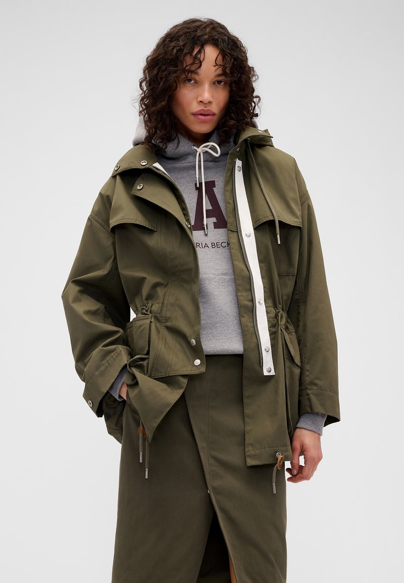 Femme aux cheveux bouclés portant une veste vert olive par-dessus un sweat à capuche gris et une jupe assortie vert olive, debout devant un fond uni.