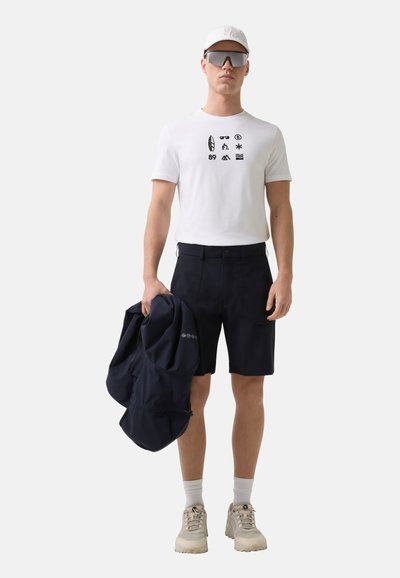 Weißes T-Shirt mit kurzen Ärmeln und schwarzen Grafiken, dunkelblaue Shorts, eine dunkelblaue Jacke tragend, Sonnenbrillen und eine weiße Kappe tragend, beigen Sneakers.