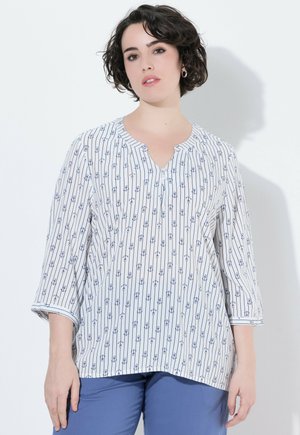 Mujer con el cabello corto, oscuro y rizado, que lleva una blusa blanca de manga larga con patrones verticales de anclas azules y pantalones azules sobre un fondo blanco.