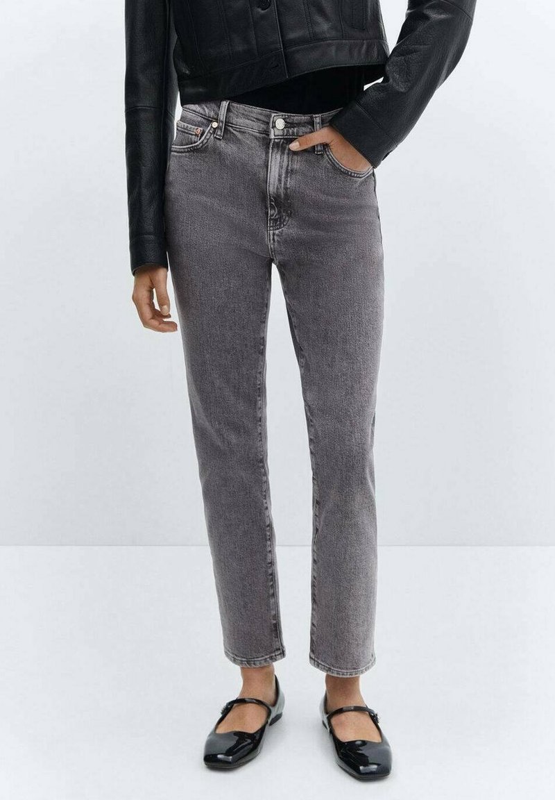 Personne portant un jean gris taille haute, une veste courte en cuir noire et des chaussures Mary Jane noires vernies, debout devant un fond uni.