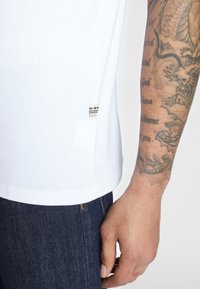 Tricou alb din bumbac cu mâneci scurte, având un mic eticheta G-Star pe tiv, asortat cu blugi din denim închis.