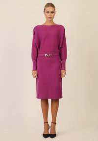 Vestido de punto fucsia con cuello barco, mangas largas, cintura ceñida que presenta un detalle de cadena plateada, de largo hasta la rodilla y con una textura suave.