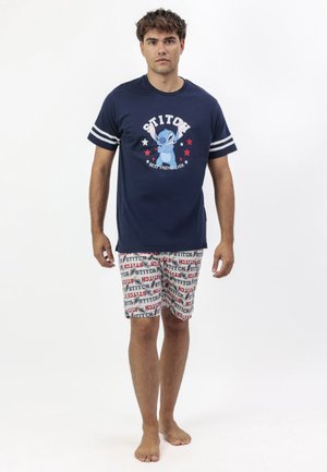 Joven de pie descalzo vistiendo una camiseta azul marino con estampado de Stitch y rayas blancas, y pantalones cortos a juego con patrones de Stitch en rojo y azul.