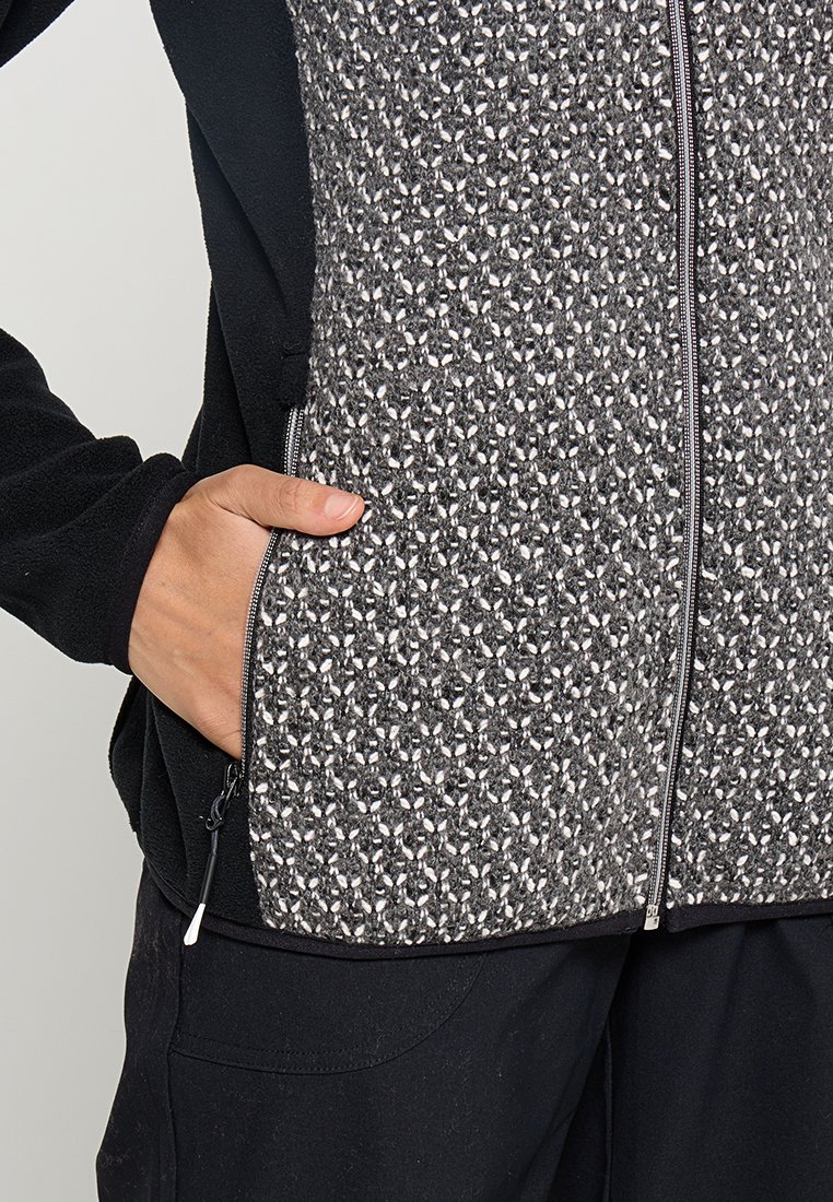 Fleecejacke mit einem strukturierten grauen und weißen Muster auf der Vorderseite, schwarzen Ärmeln und einem Reißverschluss. Seitentasche sichtbar.