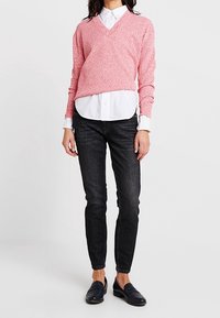 Maglione rosa a V con texture, indossato sopra una camicia bianca con bottoni, abbinato a jeans neri slim-fit e mocassini neri.