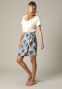 Witte V-hals T-shirt gecombineerd met een knielange, lichte rok met blauwe bloemmotieven en een strik in de taille. Sandalen maken de look af.