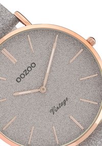 Cadran de montre argent texturé avec des accents dorés, présentant de fins index horaires, un logo à 6 heures, et un boîtier en or rose. Le bracelet est gris scintillant.