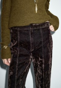Pantaloni in velluto scuro con vita alta e cuciture laterali, abbinati a un maglione texturizzato verde oliva. Dettaglio in catena dorata sul maniche.