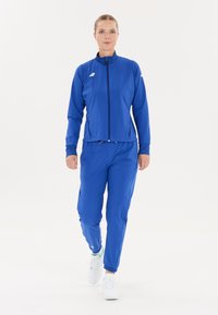 Kvinde, der går fremad iført en blå træningsdragt med lynlås-lommer og hvide løbesko mod en enkel hvid baggrund.