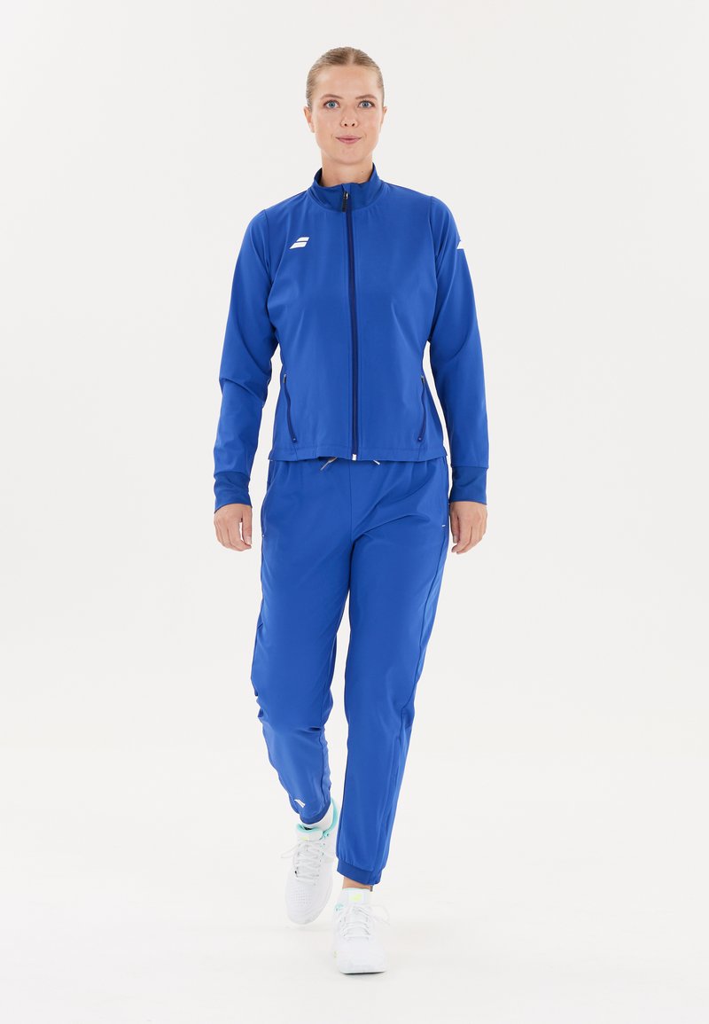 Kvinde, der går fremad iført en blå træningsdragt med lynlås-lommer og hvide løbesko mod en enkel hvid baggrund.