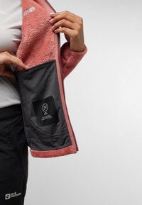 Fleecejacke mit pinkem Außenmaterial und weichem, strukturiertem Innenfutter. Verfügt über eine schwarze Innentasche mit Pflegehinweisen und Logo.