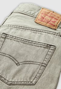 Lichtgrijze denim jeans met contrasterende donkere stiksels, een achterzak en een bruine leren patch met rode tekst en graphics.