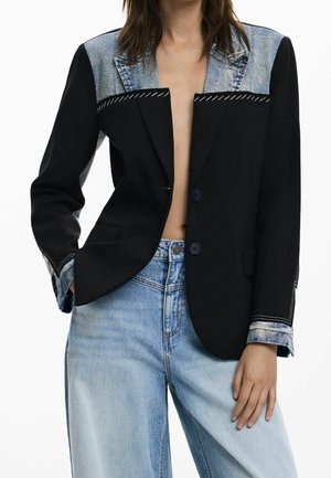 Mujer con blazer negro abierto con cuello y puños de denim combinados con vaqueros de tiro alto azul claro, mano en el bolsillo, sobre fondo blanco.