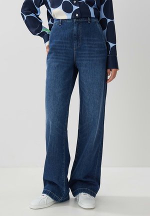 Jeans en denim bleu taille haute avec des jambes larges, présentant un design classique à cinq poches et des coutures visibles, associés à des baskets blanches.