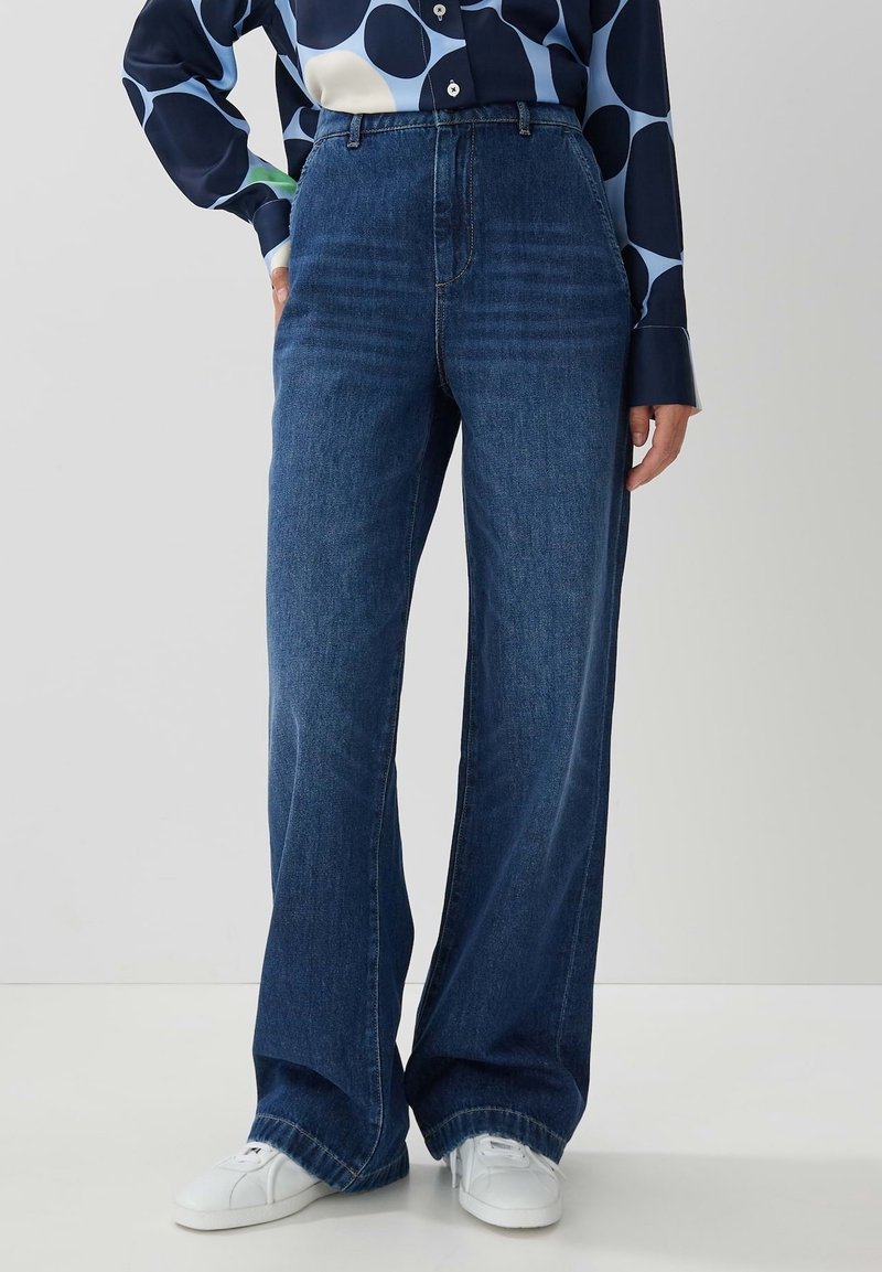 Höga blå denimjeans med vida ben, med en klassisk femficksdesign och synlig sömnad, kombinerat med vita sneakers.