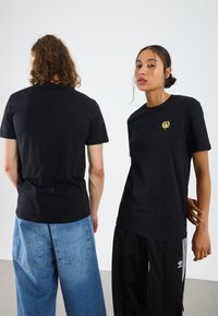 T-shirts en coton noir avec col rond ; l'un présente un petit graphique jaune. Le modèle porte un jean ample et un pantalon de sport noir.