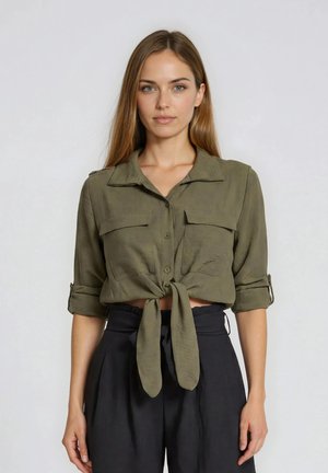Femme aux cheveux raides portant une chemise olive boutonnée nouée à la taille et un pantalon noir taille haute, debout devant un fond uni.