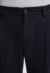 Pantaloni neri in tessuto intrecciato con cintura elastica, dotati di chiusura con bottone e un sottile motivo diagonale. Include un dettaglio con tasca frontale.