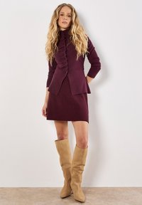 Bordeauxfarbener Rüschencardigan über einem figurbetonten Rippskleid; das Model trägt beige kniehohe Stiefel; glattes Material mit subtiler Textur.