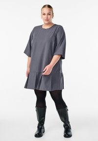 Šedé šaty ke kolenům s krátkým rukávem, sklady na spodním lemu, kulatým výstřihem, v kombinaci s černými koženými botami po kolena a tmavými punčochami.