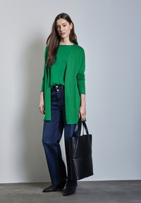 Maglietta verde a maniche lunghe abbinata a jeans in denim scuri a gamba larga. Borsa a tote nera con manici e scarpe nere a punta. Design semplice, stile minimalista.