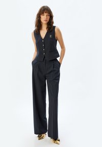 Gilet croisé bleu marine avec des rayures blanches verticales et des boutons dorés, associé à un pantalon large taille haute. Chaussures avec des accents dorés.
