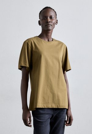 T-shirt verde oliva a maniche corte in morbido cotone con scollo tondo e vestibilità comoda, abbinato a pantaloni in denim scuro.