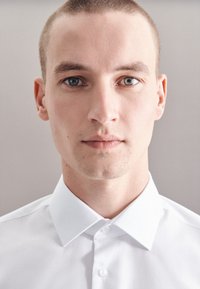 Biała koszula z klasycznym kołnierzykiem, gładkim wzorem i gładką fakturą. Model ma krótkie włosy, a tło jest neutralne.