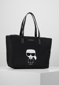 Svart nylon shoppingväska med dubbla svarta läderhandtag, som har en vit tecknad karaktärsgrafik på framsidan och en dragkedjestängning.