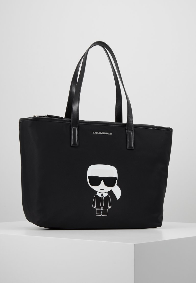 Svart nylon shoppingväska med dubbla svarta läderhandtag, som har en vit tecknad karaktärsgrafik på framsidan och en dragkedjestängning.