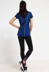 Haut de sport marine avec des bordures roses, associé à un legging noir avec un motif floral rose au niveau de la cheville, et des chaussures de sport noires.