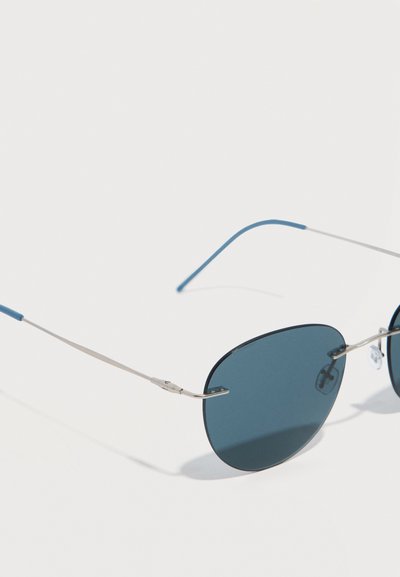 Lunettes de soleil à monture argentée avec des branches fines et des verres ovales teintés en bleu, présentées sur un fond blanc uni.